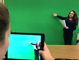 Student giving a presentation in front of a green screen as another student films using a tablet